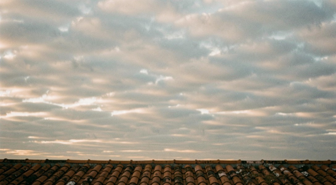 Un ciel couvert de nuages en couches au-dessus d’un toit de tuiles, lumière diffuse de fin d’après-midi, détails de la couverture nuageuse visibles, plan large.